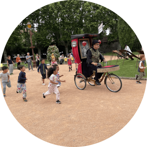 jean bobine, le magicien bonimenteur de la grande course des magiciens sur leur drôles de machines pilote son tricycle accompagné par les enfants qui courent à ses côtés